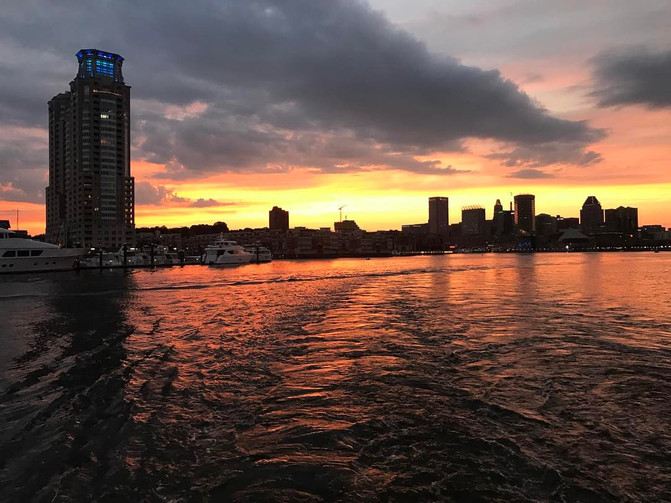 An overhead flat-lay photograph of a coastal city skyline at sunset viewed from water, with boats, a tall building, and an orange and purple sky reflected on calm waves.